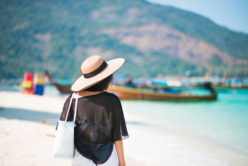 The back of one Asian woman wearing a shirt and a hat with holding bag walks on a sunny beach with blue sea and boats in the background. Concept of holidays and vacations. Picture looking alone or sad
