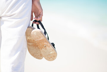 Close up a hand with tan skin holding slippers on the sunny beach and seaside background in the...