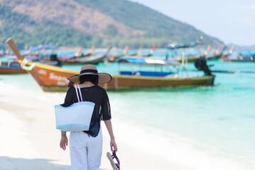 The back of one Asian woman wearing a shirt and a hat with holding bag walks on a sunny beach with blue sea and boats in the background. Concept of holidays and vacations. Picture looking alone or sad