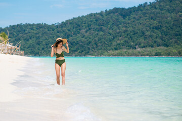 Obraz premium Long shot Asian women smiling wearing and holding straw hat and green bikini enjoy on beach with seaside and resort background. Concept of happy holidays and relaxing vacations. Copy space for text.