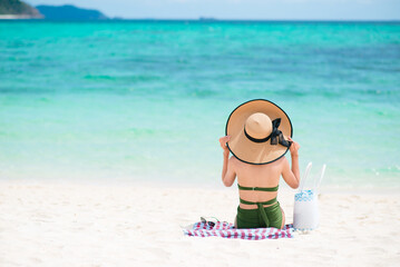 Back of an Asian woman wearing and holding straw hat and green bikini sitting on beach with blue sea and sky background in the daytime. Concept of happy holidays and relax travel. Copy space for text.
