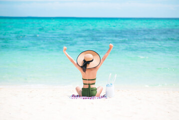Back of an Asian woman wearing straw hat and green bikini outstretched arm sitting on beach with blue sea and sky background in the daytime. Concept of happy holidays and travel. Copy space for text.