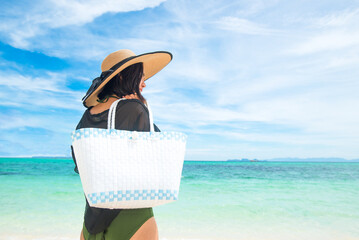 Back of Asian woman wearing See-through and green bikini holding a bag on a sunny beach with sea and sky background in the daytime. Concept of summer holidays and travel. Copy space for text.