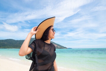 Asian woman wearing sunglasses, hat, See-through, and bikini. hand holding a hat on the beach with clear sea and sky background. long weekend holidays and travel concept. Relaxed. Copy space for text.