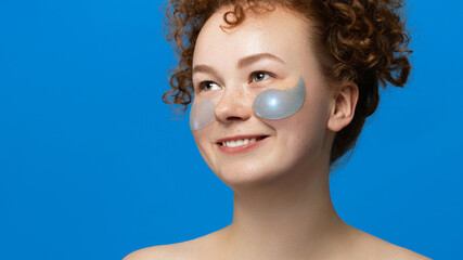 Smiling young woman with curly hair and freckles poses with silver under-eye patches. Her fresh...