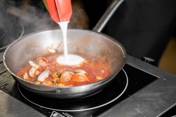 A chef adds cream to a skillet filled with simmering clams in a rich tomato sauce, creating a delightful cooking scene with steam rising on a stovetop.