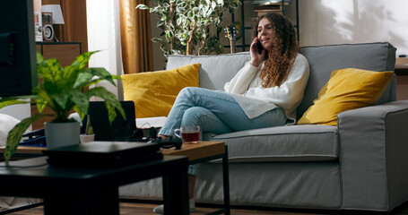 Fototapeta premium Happy woman sits on the couch, turns on a movie, and talks to a friend on the phone. She laughs, gossips, and enjoys the moment while the TV plays in the background.