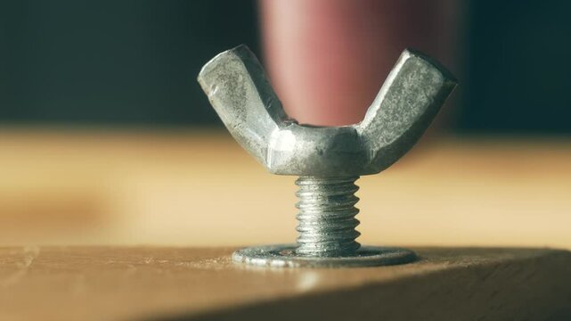 Close-up metal wing nut screwing onto bolt by man using hands. Macro view hardware fastener tightening manually without tool by repairman. Steel nut working onto thread by builder with fingers during
