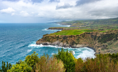Miradouro de Santa Iria, Ribeira Grande, São Miguel, Azoren: Panoramablick auf die schroffe Nordküste und üppige Flora. Blaues Meer, grüne Landschaft und bewölkter Himmel schaffen eine träumerische At