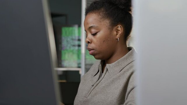 Medium shot of focused young African American female software developer sitting in front of PC at office desk with running program codes on screen in blurred background