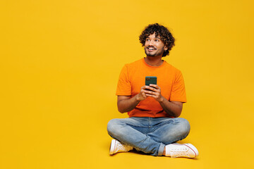 Full body young Indian man wear orange t-shirt casual clothes sit hold in hand use mobile cell...