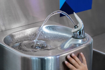 A child touches the handle of a modern drinking fountain as water flows in an indoor setting, emphasizing hydration and accessibility. The stainless steel surface is slightly wet.