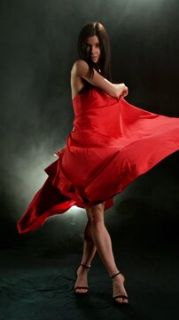 Stunning Black-Haired Woman in Ruby Red Dress and High Heels Gracefully Posing Against a Black Background, Captured in Super Slow Motion. Vertical Aspect Ratio. Speed Ramp Effect.