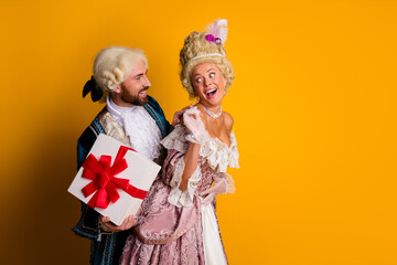 Couple in historical costumes sharing a joyful moment with a gift against a vibrant yellow...
