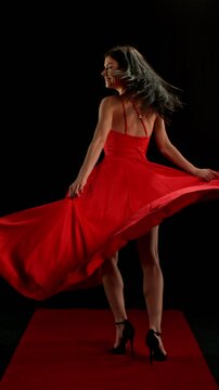 Stunning Black-Haired Woman in Ruby Red Dress and High Heels Gracefully Posing Against a Black Background, Captured in Super Slow Motion. Vertical Aspect Ratio.