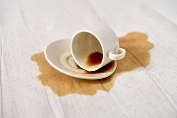 Empty coffee cup tipped on white tablecloth, creating brown liquid stain. Soft lighting accentuates stark contrast between white fabric and dark spill, conveying sense of accident or clumsiness