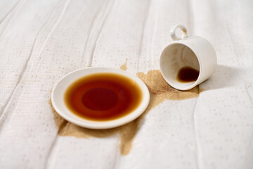 Coffee cup tipped over on light beige fabric. Dark coffee forms pool under saucer. Soft, diffused lighting emphasizes fluids deep hue