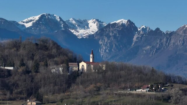 A beautiful aerial telephoto shot with a smooth left-to-right tracking movement capturing the Church of Bolca, nestled in the hills of Verona, with the majestic Monte Carega as a backdrop.