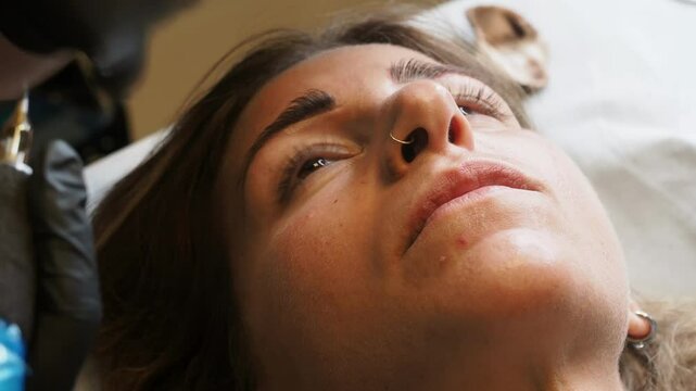 Slow motion close-up of eyebrow microblading with gloved hands and blue cosmetic tool on relaxed client