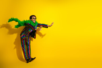 Young stylish man in vibrant striped suit and green boa posing against yellow background,...