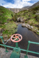 A vibrant green landscape showcases a rustic valve set beside a serene stream, which flows towards a distant Silent Valley Reservoir dam nestled in Mourn Mountains, Northern Ireland