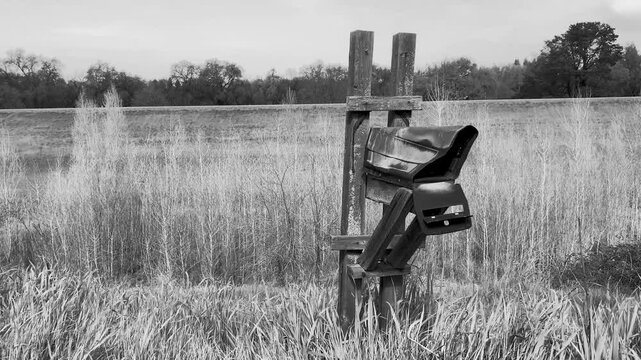 Grayscale of rusty and damaged mailbox in a rural field with tree line in the background