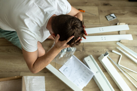 man clutches his head looking at furniture assembly diagram. man tries to assemble complex furniture by himself