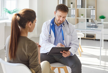 Obraz premium Male doctor checking female patient symptoms, holding using clipboard, listening to woman complain, sitting in hospital office during medical checkup and healthcare, physician gp consulting client