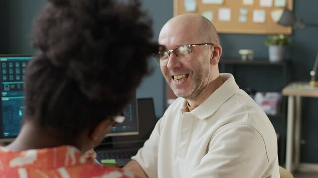 Medium close up shot of friendly middle-aged Caucasian male employee chatting and giving high five to young African American female coworker sitting at one desk in open space office in front of PC