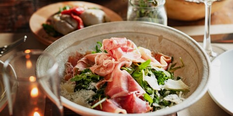 A fresh salad with prosciutto, arugula, and parmesan in a bowl. The salad is garnished with herbs, creating a vibrant and appetizing dish on a dining table. Aragula salad with parma ham, prosciutto.
