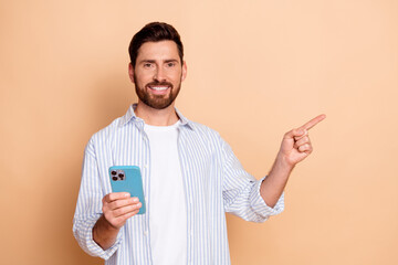 Smiling young man holding smartphone in striped shirt pointing to the side with confident gesture...