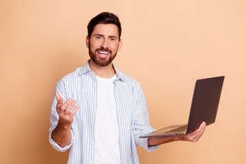 Young man holding laptop in a casual striped shirt with a confident expression against a beige background, showcasing a cheerful demeanor
