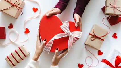Hands exchanging a heart-shaped gift box with ribbon, surrounded by wrapped presents and red hearts. Gift exchange, heart-shaped box, and presents theme. Gift exchange for Valentines or festive events