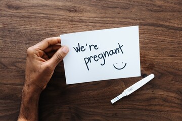 A hand holds a card with 'We're pregnant' and a smiley face, next to a pregnancy test on a wooden table. Pregnancy announcement with a positive test. Man surprised by pregnancy announcement on card.