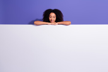 Portrait of a stylish young woman with curly hair peeking over a blank board against a purple...