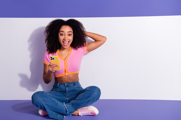 Young woman with curly hair holding a phone, sitting cross-legged on a purple and white background