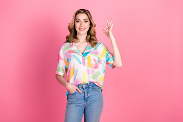 Charming young woman in colorful shirt smiling and gesturing OK on pink background