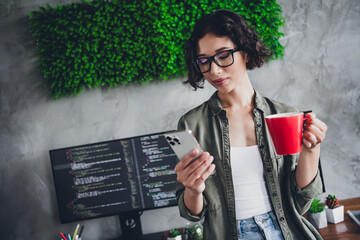 Young female programmer checking her smartphone while holding a coffee mug in her modern home...