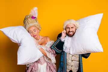 Amusing Historical Couple Dressed in Royal Costumes, Holding Pillows Against a Vibrant Yellow...