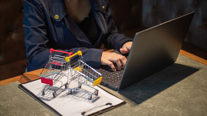 Businesswoman working on laptop about selling products. In front of her are documents and a small cart. Online trading and shopping.