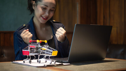 Businesswoman smiling and gesturing happily at successful online deal closing via laptop and shopping cart conveying trade.