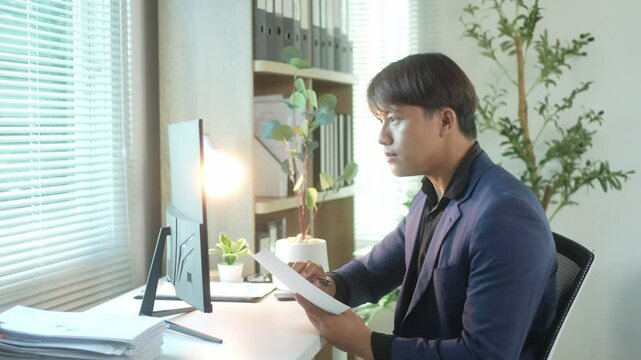 A man in a suit is sitting at a desk with a computer monitor and a stack of papers. He is writing on a piece of paper with a pen. The scene suggests a professional work environment