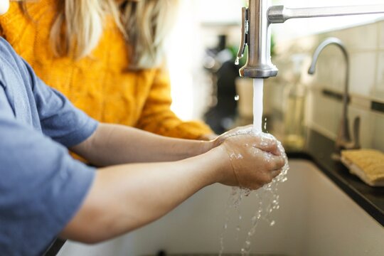 Child washing hands under faucet, water flowing, hygiene focus. Woman watching the boy washing hands at sink for hygiene and cleanliness. Boy washing his hands with the help of his mother at home