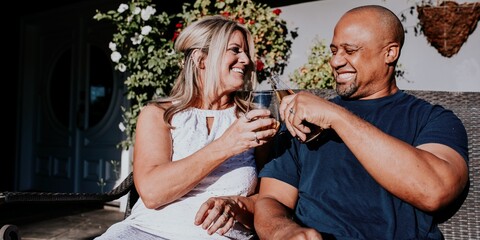 A couple enjoying drinks outdoors, smiling and toasting. Relaxed atmosphere with greenery. Happy couple, outdoor drinks, and smiles. Couple sitting together at night, diverse happy couple.