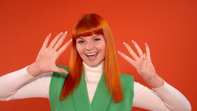 Cheerful young woman with vibrant orange hair and a green vest, smiling and showing nine 9 fingers against a bright orange background.