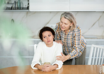 Kind old woman asking for forgiveness to offended middle-aged woman sitting in the kitchen