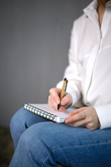 In the photo, a woman in a white shirt and blue jeans sits writing in a notebook with a pen. He is focused on his work, his hand resting on the notepad. The background is a blurred gray wall