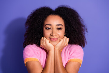 Smiling young woman with curly hair wearing a pink fashion sweater against a purple background...