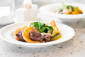 A gourmet dish featuring tender beef, crispy potatoes, and fresh vegetables is elegantly plated on a white dish, with a second pasta plate blurred in the background, highlighting culinary expertise.