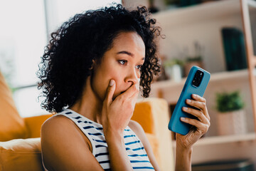 Young woman with curly hair expressing surprise while looking at smartphone in cozy modern home...